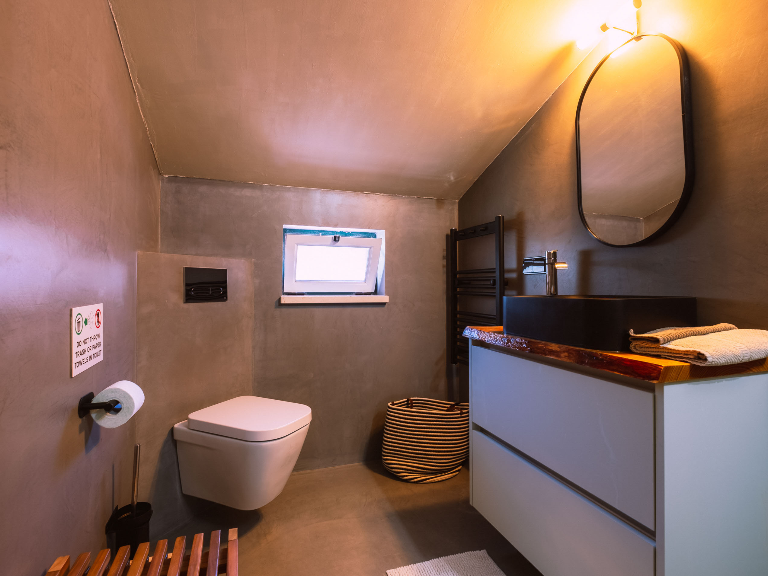 Bathroom with wall-mounted toilet, small window, sink with mirror, striped laundry basket, and towel warmer.